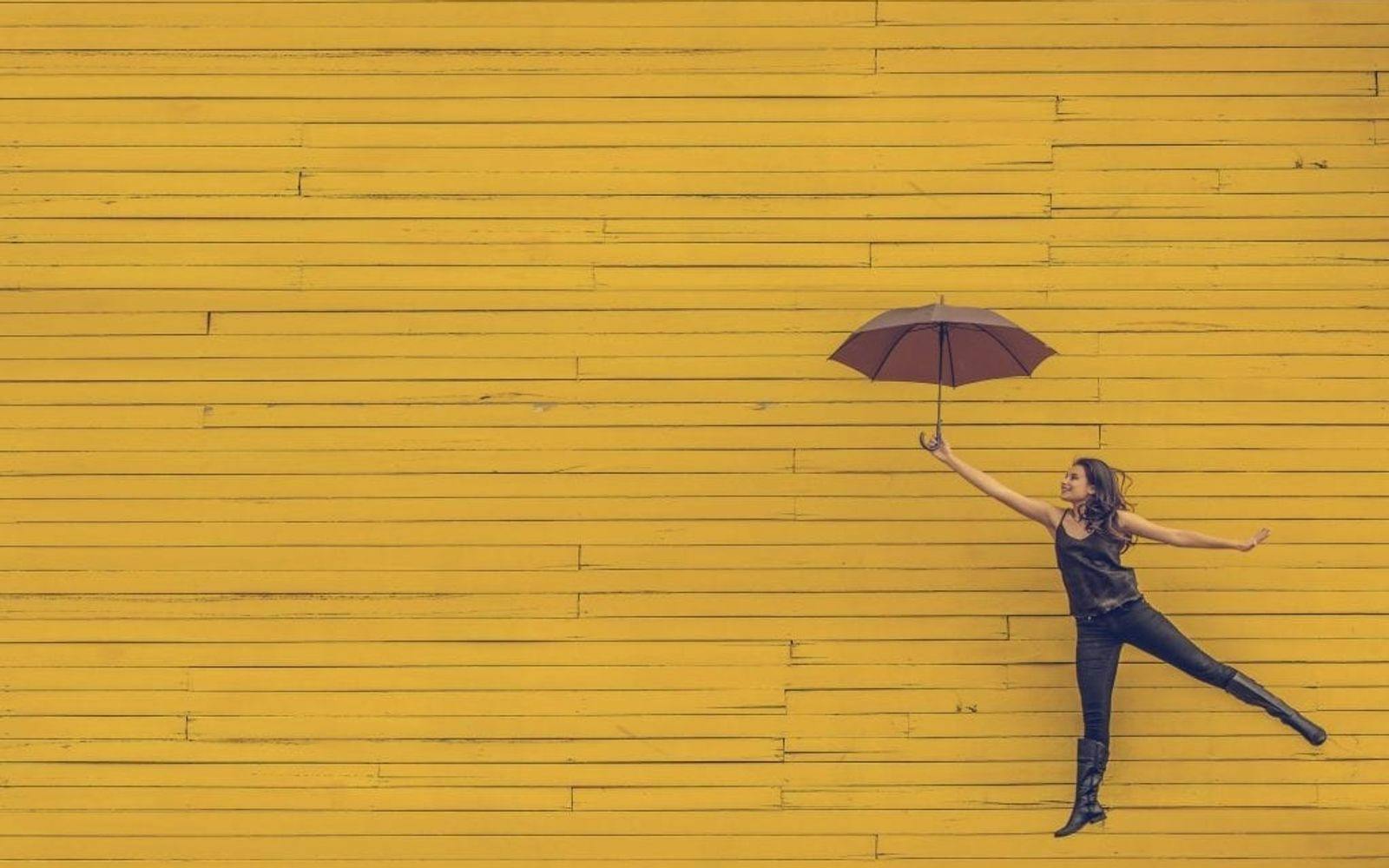 Kvinne foran gul bakgrunn som flyr med en paraply som Mary Poppins inn i bildet