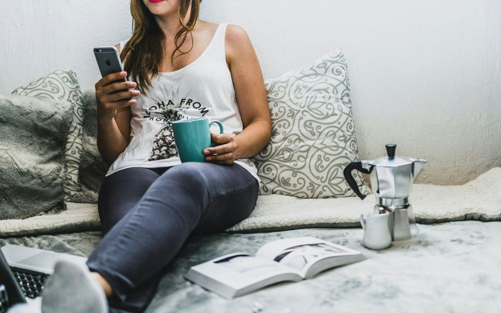 Kvinne som sitter avslappet med kaffekopp og jobber på mobilen